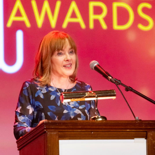 Brunette lady standing in front of a brown podium in front of red Staff Excellence Awards backdrop